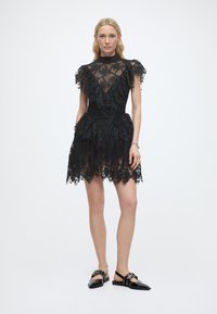 Γυναίκα που φοράει μαύρο δαντελένιο φόρεμα με στρώσεις βολάν και διαφανή μανίκια, συνδυασμένο με μαύρες μυτερές μπαλαρίνες με λουριά και λεπτομέρειες αγκράφας.