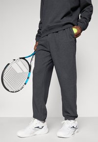Person i mørkegrå joggebukser og genser som holder en tennisracket og en gul tennisball, iført hvite joggesko.