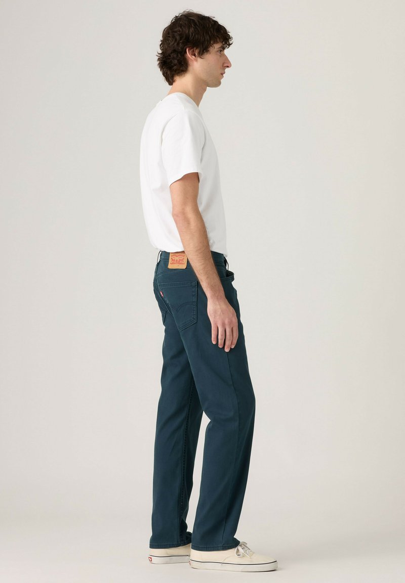 Mann steht seitlich, trägt ein weißes T-Shirt, dunkelblaue Jeans mit einem Lederpatch und weiße Sneaker vor einfachem Hintergrund.