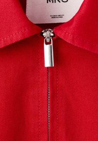 Prim-plan cu guler de îmbrăcăminte roșie cu fermoar argintiu, arătând cusăturile și o etichetă de îmbrăcăminte parțial vizibilă.