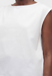 Gros plan d'une personne portant un haut blanc sans manches avec un col bateau, montrant le haut du torse et le bras droit.