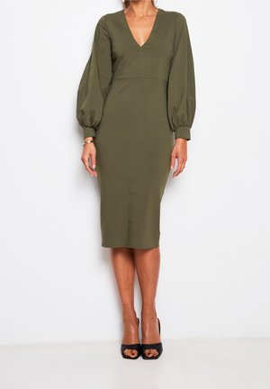 Nainen pukeutuneena oliivinvihreään puolipitkään mekkoon, jossa on pitkät liehuvat hihat, V-aukko ja mustat avoimet korkokengät, valkoista taustaa vasten.