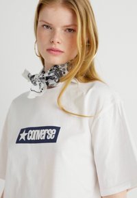 Weißes Baumwoll-T-Shirt mit einem navyblauen "Converse"-Logo und Stern-Grafik auf der Vorderseite. Modell trägt einen gemusterten schwarz-weißen Schal.