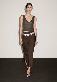 Pantalons larges en satin marron avec une ceinture métallique accentuée et un débardeur en tricot foncé. L'ensemble présente un design minimaliste et contemporain.
