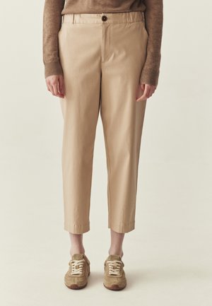 Osoba w beżowych, krótkich spodniach, brązowym swetrze z długim rękawem i beżowych trampkach, stojąca na tle jednolitego, jasnego tła.