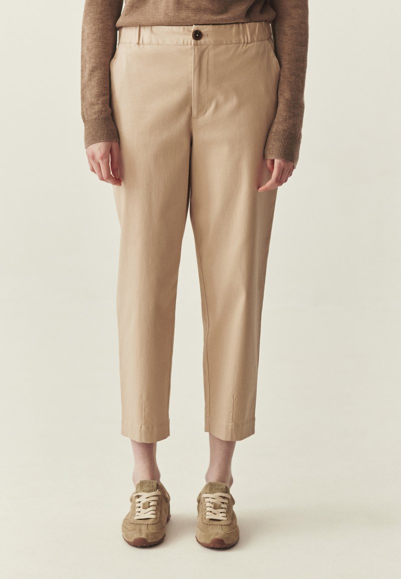 Osoba w beżowych, krótkich spodniach, brązowym swetrze z długim rękawem i beżowych trampkach, stojąca na tle jednolitego, jasnego tła.