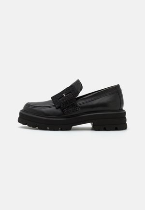 Mocassin en cuir noir avec une semelle épaisse et une large sangle texturée avec boucle sur la partie supérieure, présenté de profil sur fond blanc.