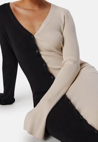 Femme portant une robe côtelée boutonnée avec un côté noir et l'autre beige, assise avec les bras croisés sur les genoux.