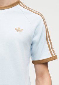 Ανοιχτό μπλε κοντομάνικο πουκάμισο με καφέ λεπτομέρειες στη γιακά, τις άκρες των μανικιών και ρίγες στους ώμους, με μικρό καφέ λογότυπο Adidas στο στήθος.