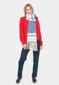 Naine, kes kannab punast kampsunit, siniseid teksaseid, pruune kingi ja pikkade siniste ja valgete triibuliste narmastega salliga, seisab ühevärvilise tausta ees.