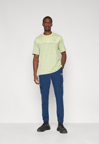 Ανοιχτό πράσινο αθλητικό μπλουζάκι με κοντά μανίκια και υφασμάτινο σχέδιο, συνδυασμένο με σκούρο μπλε παντελόνι εργασίας με τσέπες και λεπτομέρειες.