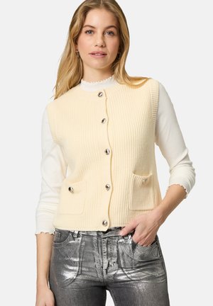 Cream ribbed knit vest with silver buttons and two front pockets, worn over a white top. Paired with metallic silver jeans.
