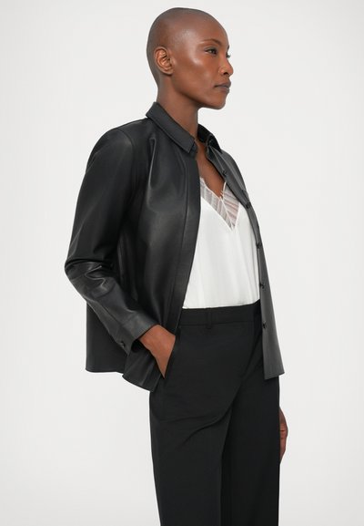 Must have musta nahktagi sujuva tekstuuriga ja nööpidega, kombineeritud valge pluusi ja mustade pükstega, kujutatud külgprofelis.