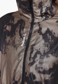 Reebok zipjacka i strukturerat svart och guldmönster, med hög krage och logotypaccent på framsidan.