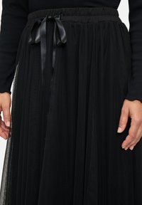 Gonna in tulle nera con un elastico in vita e un nastro in satin nero. Il tessuto presenta uno strato trasparente e testurizzato con pieghe verticali.