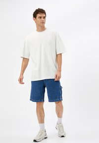 Camiseta de algodón blanca de gran tamaño combinada con shorts de mezclilla azul, con un corte holgado y bolsillos laterales. Llevada con zapatillas blancas y calcetas de canalé.
