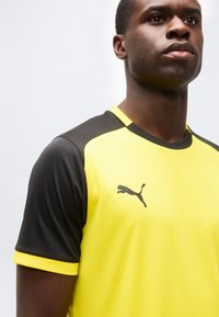 Άνδρας που φοράει αθλητική μπλούζα κίτρινου και μαύρου χρώματος με μαύρο λογότυπο Puma στο στήθος, εμφανιζόμενος από τους ώμους και πάνω μπροστά σε ένα απλό φόντο.