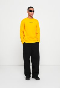Yellow sweatshirt with black text, paired with loose-fitting black cargo pants and black sneakers. The model wears dark sunglasses.