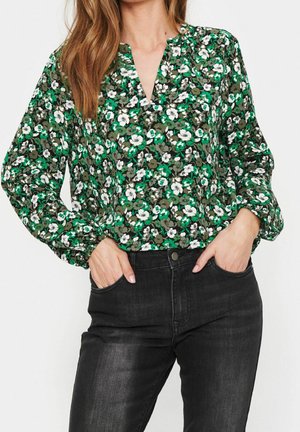 Vrouw draagt een blouse met groene, witte en zwarte bloemen, lange mouwen en een V-hals, gestopt in een zwarte jeans met hoge taille, handen in de zakken.