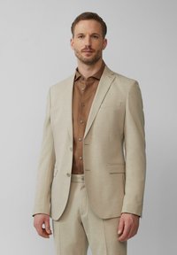 Lichtbeige pak met een getextureerde weving, gecombineerd met een bruine geknoopte blouse. Opvallende details zijn onder andere twee knopen op de blazer en een borstzak.