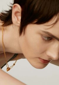 Collar de oro con un colgante de corazón abstracto, que presenta una delicada cadena y detalles texturizados, llevado por un modelo con cabello corto.