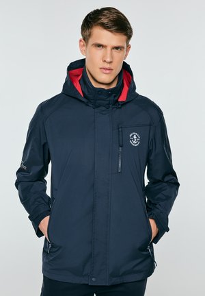 Młody mężczyzna w granatowej kurtce z kapturem i czerwoną podszewką, stojący z rękami w kieszeniach, na neutralnym szarym tle.