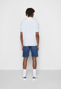 Camisa de manga corta con cuello y rayas verticales azules y blancas, combinada con pantalones cortos de mezclilla azul. El conjunto se completa con calcetines blancos y zapatillas deportivas.