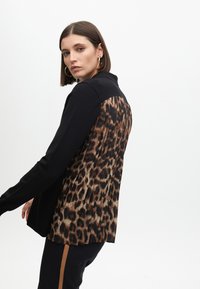 Černý dlouhý rukáv top s skládáním na hnědém leopardím potisku na zadní straně; má límec a hladkou přední část z látky.