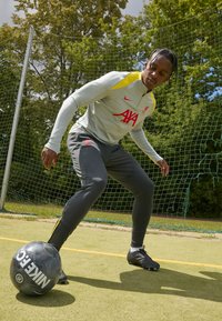 Camiseta gris de manga larga con detalles amarillos y el logo del Liverpool, pantalones grises y botas negras. El jugador está driblando un balón de fútbol Nike en blanco y negro.