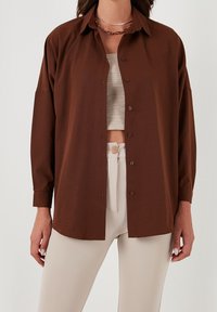 Donna che indossa una camicia marrone larga con bottoni sopra un top a canotta beige a coste e pantaloni beigi alti in vita con un bottone di legno.
