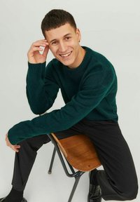 Pull en laine vert avec des poignets côtelés, associé à un pantalon noir et des chaussures, assis sur une chaise en bois dans une pose détendue.