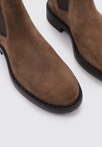 Botas de tobillo de ante marrón con punteras redondeadas y paneles laterales elásticos; costuras en contraste a lo largo de la suela y un tacón plano negro.