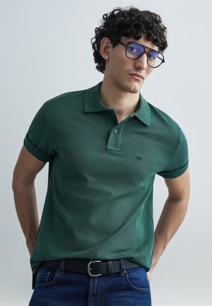 Zielona koszulka polo z krótkimi rękawami, wykonana z teksturowanej tkaniny, z kołnierzykiem na dwa guziki oraz małym haftowanym logo na piersi, zestawiona z niebieskimi dżinsami i czarnym paskiem.