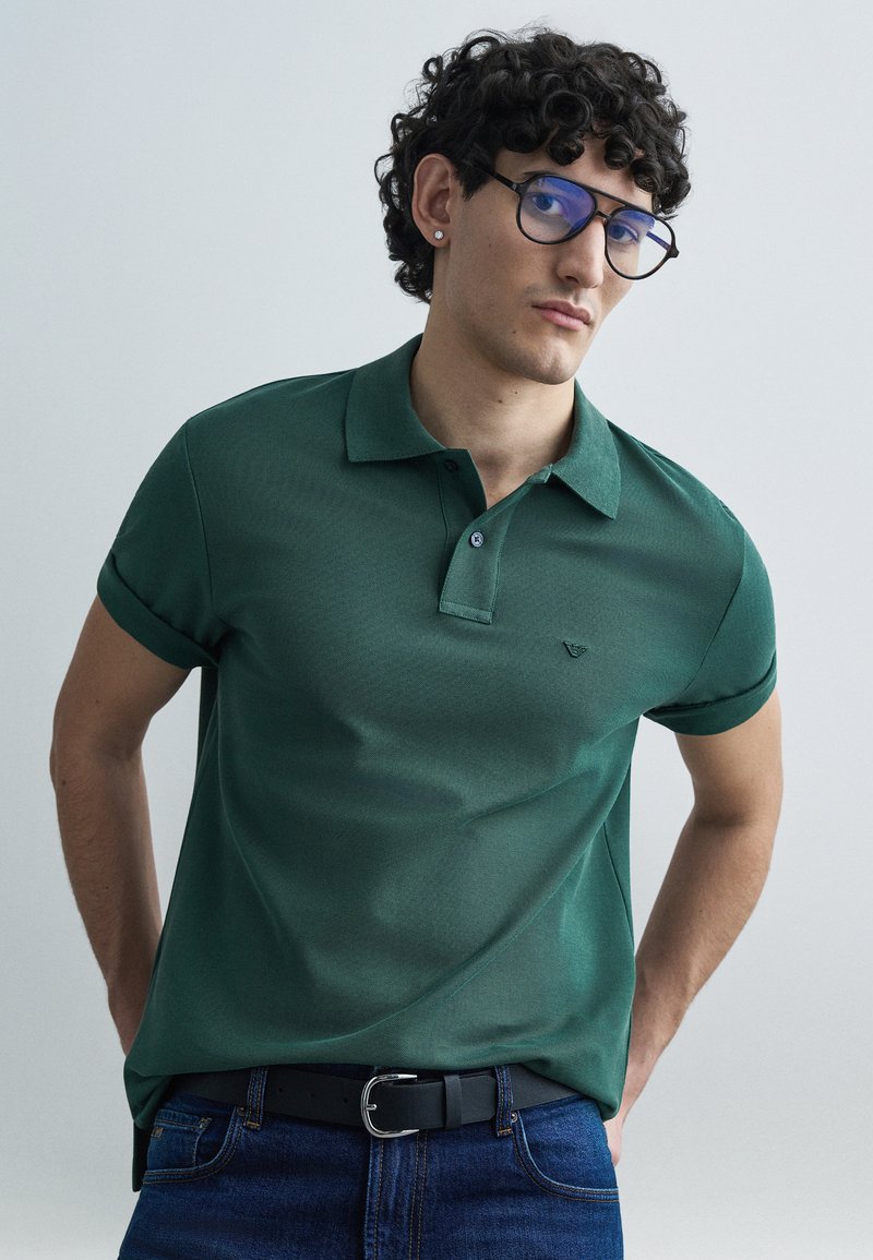 Polo vert à manches courtes, tissu texturé, col à deux boutons, et petit logo brodé sur la poitrine, assorti à un jean bleu et une ceinture noire.