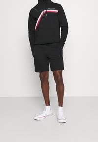 Schwarzer Kapuzenpullover mit einem grafischen Streifen in Rot, Weiß und Blau, kombiniert mit schwarzen Shorts und weißen High-Top-Sneakers.
