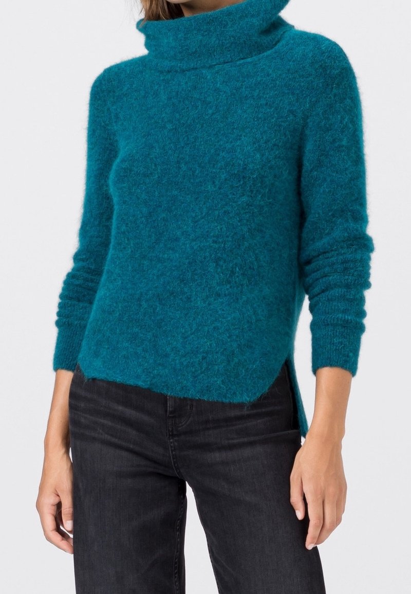 Femme portant un pull à col roulé duveteux de couleur bleu sarcelle à manches longues et un jean noir, posant devant un fond blanc uni.