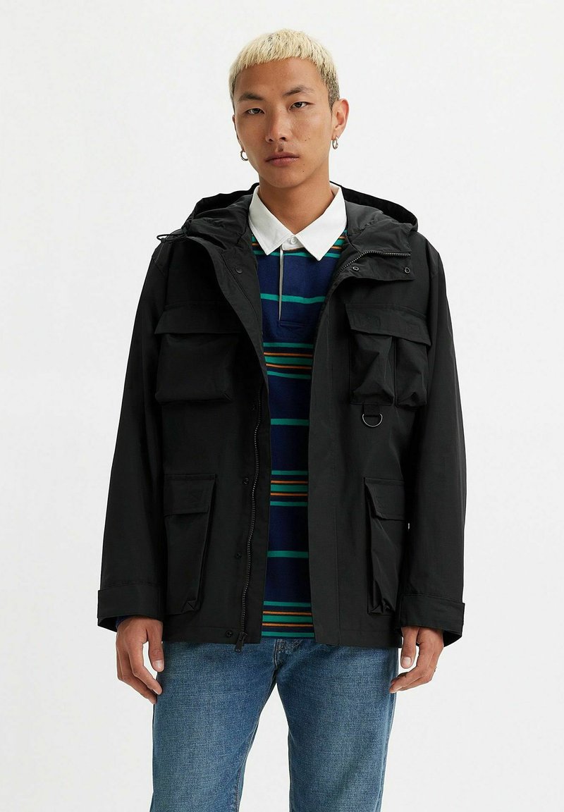 Veste noire à capuche avec plusieurs poches avant et une fermeture éclair. Portée par-dessus une chemise rayée bleue et verte, associée à un jean bleu.