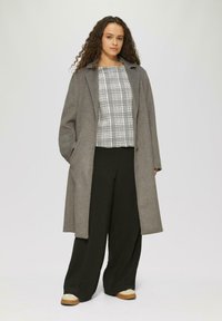 Grauer Wollmantel mit lässigem Schnitt, getragen über einer schwarz-weißen karierten Bluse, kombiniert mit schwarzen weiten Hosen und beigefarbenen Schuhen.