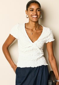 Haut blanc texturé avec un col en V et des manches courtes ; présente un design froncé et un ourlet volanté. Associé à un pantalon bleu marine et un sac noir.