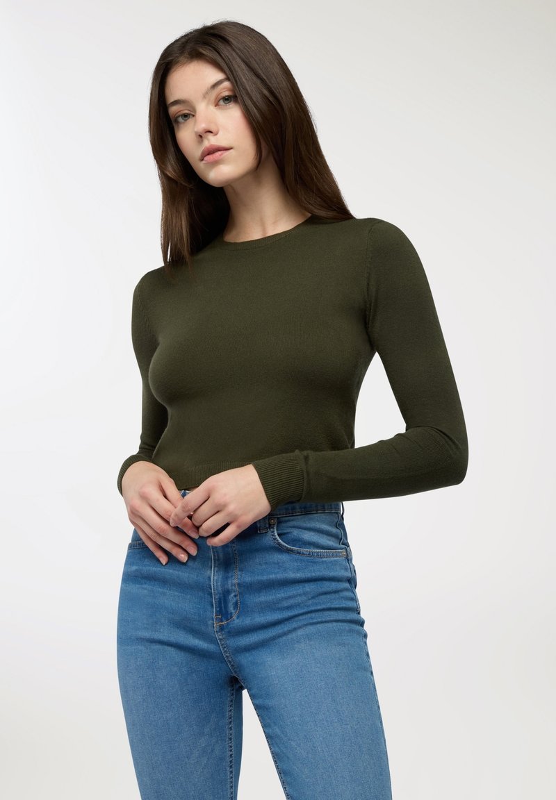 Oliwkowa bluzka z długim rękawem i okrągłym dekoltem, z ściągaczami na rękawach, połączona z niebieskimi dżinsami o wysokim stanie. Gładka faktura.