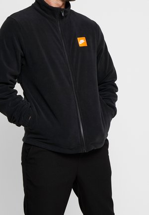 Mężczyzna w czarnej polarowej kurtce na zamek błyskawiczny z pomarańczowym logo Nike na piersi, ręce w kieszeniach, na jednolitym jasnym tle.