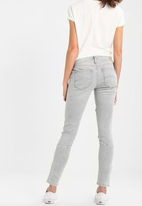Mujer vistiendo jeans ajustados de color gris claro, camiseta blanca de manga corta y zapatillas blancas, de pie de espaldas sobre un fondo blanco.