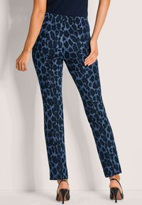 Vrouw in slanke blauwe en zwarte dierenprintbroek met een zwart topje en zwarte hoge hakken, staand tegen een lichte muur.