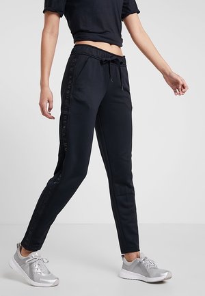 Personne portant un pantalon de jogging noir avec le texte « Under Armour » sur les côtés, un haut court noir et des chaussures de sport grises, debout sur un sol gris.
