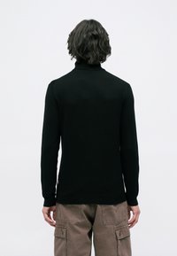 Pessoa com cabelo ondulado escuro, vestindo uma blusa preta de manga longa e calças cargo castanhas, de pé de costas contra um fundo claro.