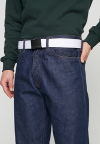 Dunkelblaue Denim-Jeans mit lockerer Passform, mit orangen Nähten und einem weißen elastischen Gürtel mit einer schwarzen Metallschnalle.