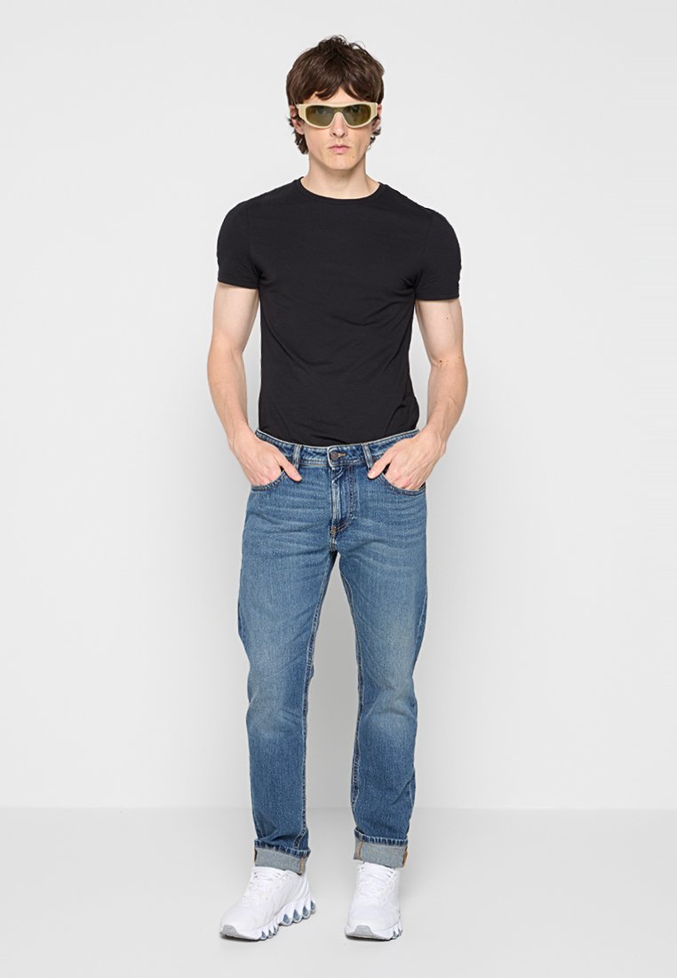 Jeune homme portant des lunettes de soleil jaunes, un t-shirt noir, un jean bleu retroussé et des baskets blanches, se tenant avec les mains dans les poches.