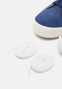 Blå mockaskor med vit gummisula, ihop med två runda, vita bomullssnören med plastspetsar.