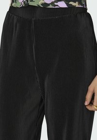 Juodos spalvos klostuotos kelnės su tekstūruota paviršiaus struktūra ir elastinga juosmens juosta. Audinys yra lygus, puoštas vertikaliomis juostomis, kurios sustiprina dizainą.