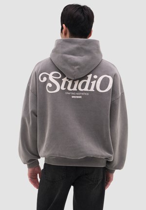 Persoon die een grijze capuchontrui draagt met de tekst "Studio" en "Crafting Aesthetics WRSTBHVR" op de achterkant, staand met de rug naar de kijker.